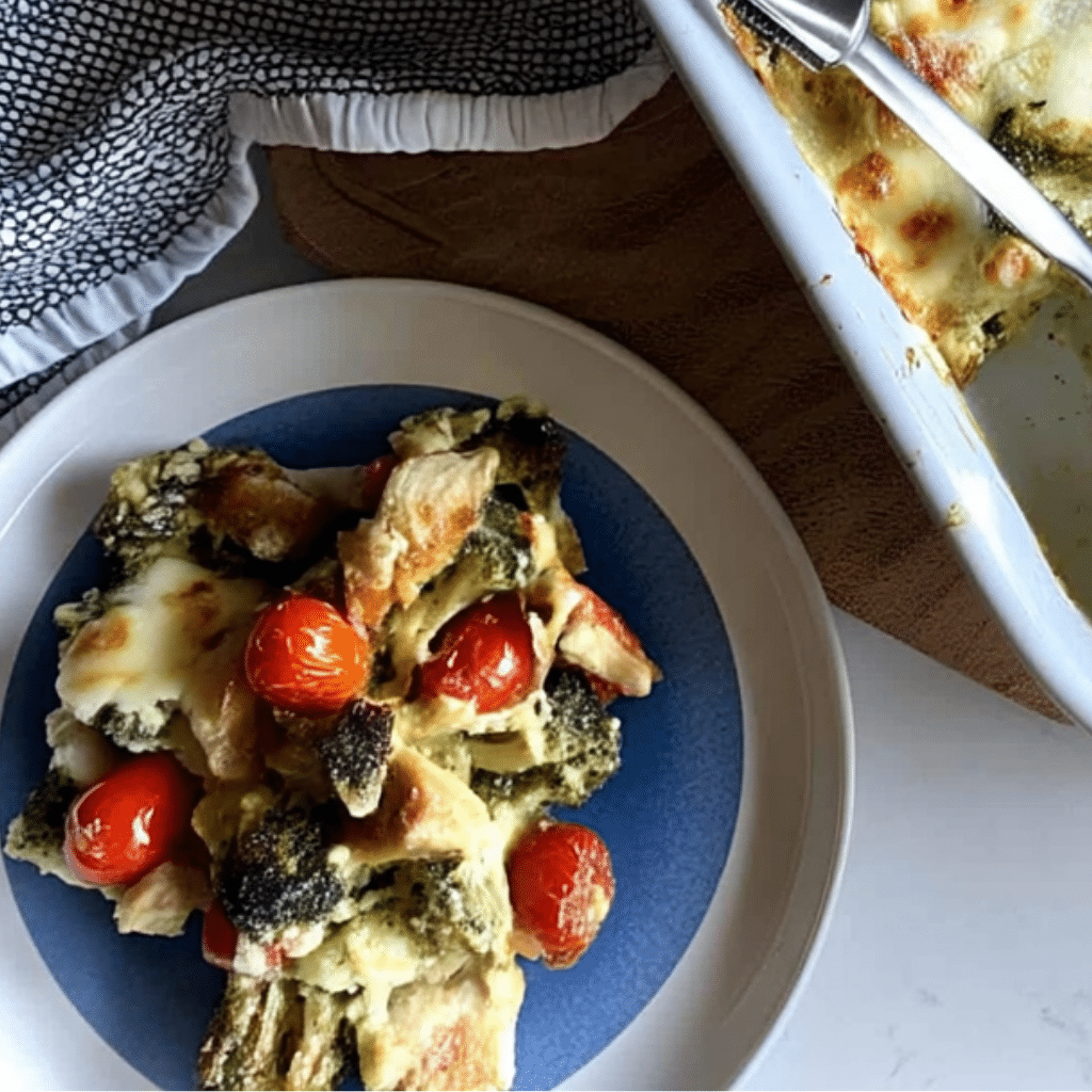 Overhead view of a blue-centered plate heaped with Tuscan Chicken Broccoli Bake (chicken, broccoli, cherry tomatoes, melted cheese), next to a partly visible white baking dish and a black-and-white patterned napkin.